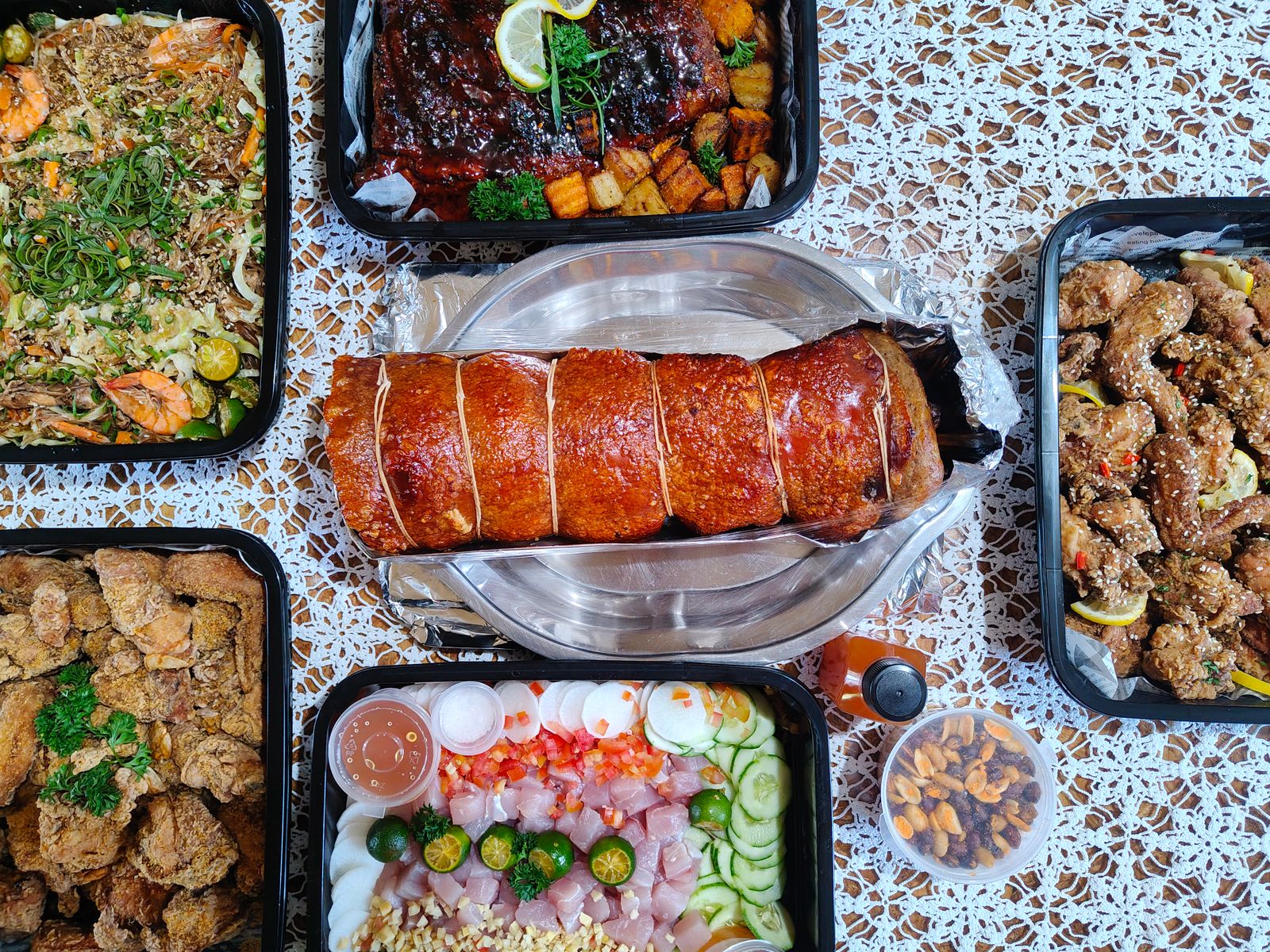 A Filipino feast spread on a lace tablecloth with a whole lechon belly roll at the center