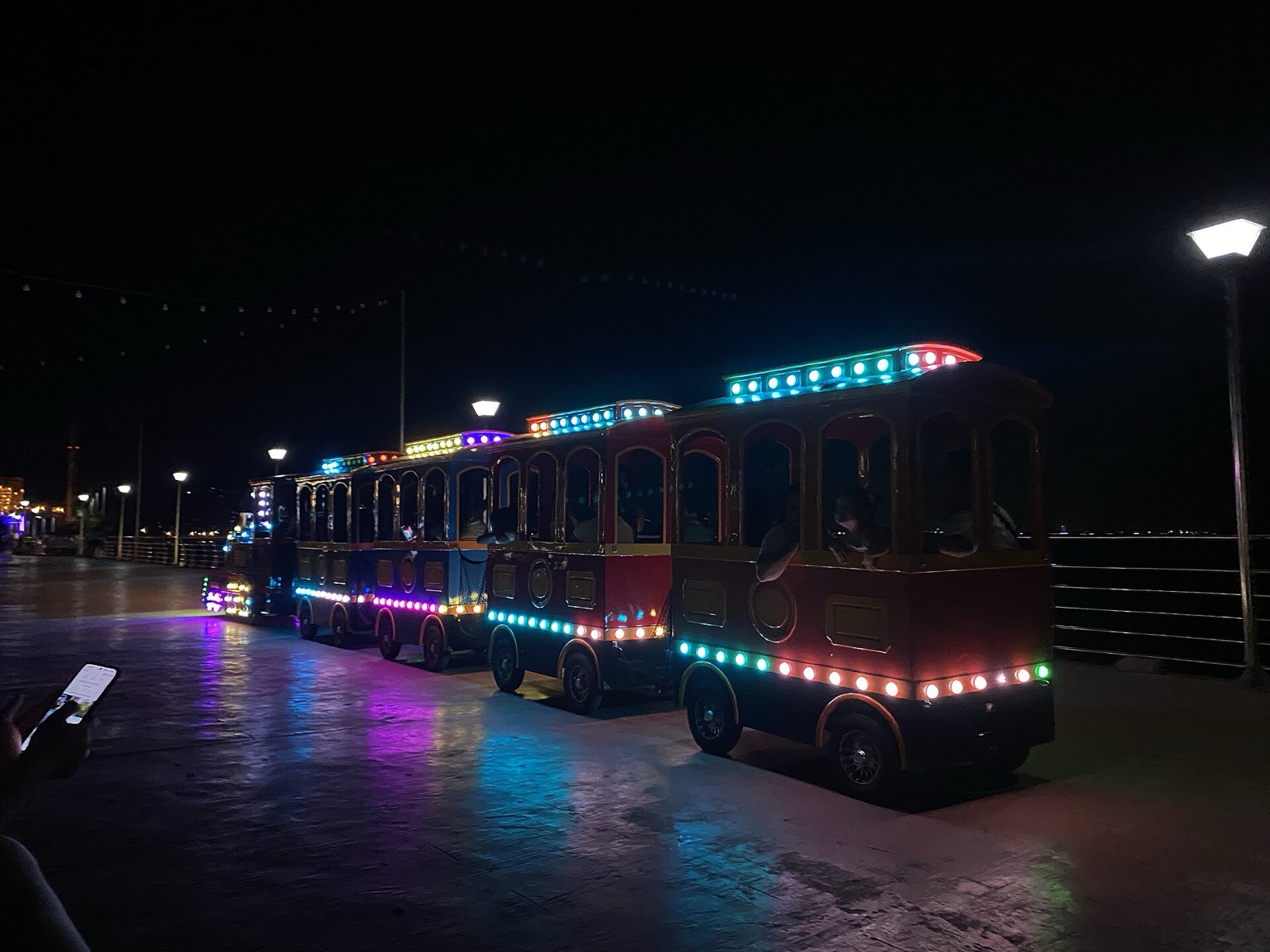 A small trolley train covered in colored lights rolling along the Naga boardwalk at night